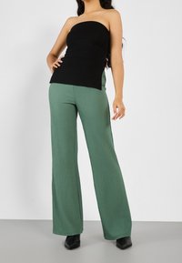 Černý top bez ramínek s bočním nabíráním, doplněný zelenými širokými ribbed kalhotami. Černé boty na špičce dokončují celý outfit.