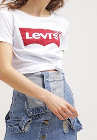 Mujer vestida con una camiseta blanca de Levi's atada a la cintura y una falda de mezclilla desgastada con tirantes y botones.