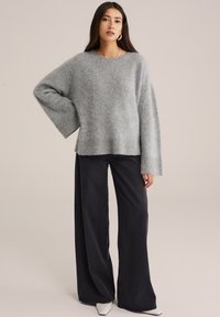 Szary oversize sweter z dzianiny z szerokimi rękawami i ściągaczami, połączony z czarnymi spodniami o wysokim stanie i szerokich nogawkach oraz spiczastymi białymi butami.