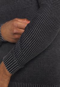 Czarny sweter o teksturowanej fakturze z prążkowanym wzorem, z wyeksponowaną ręką wsuniętą w rękaw, ukazującą elastyczne mankiety i detale materiału.