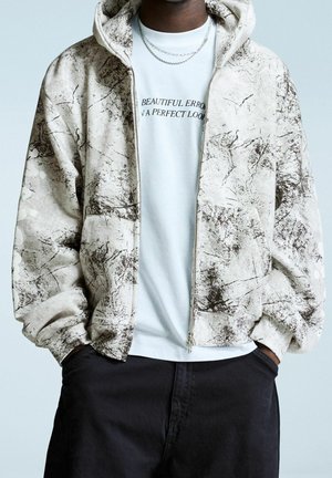 Persona que lleva una sudadera blanca con cremallera y un patrón abstracto negro, camiseta blanca con el texto "Beautiful error a perfect look", pantalones negros y cadenas plateadas.
