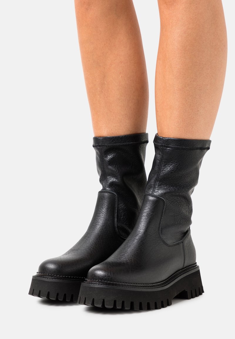 Bottes en cuir noir mi-mollet avec semelles crantées épaisses, portées sur des jambes nues sur un fond clair uni.
