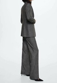 Veste grise cintrée avec pantalon large assorti. Confectionnée en tissu texturé, elle présente des épaules ajustées et une silhouette longue. Chaussures noires à bouts pointus.