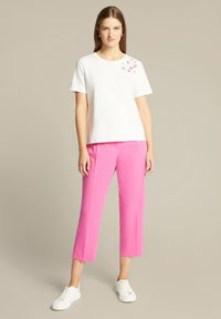 Witte t-shirt met bloemenborduurwerk op de linkerschouder, gecombineerd met felroze enkellange broek en witte sneakers.