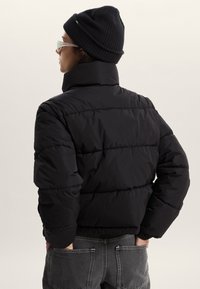 Giacca imbottita nera con collo alto e motivo trapuntato orizzontale. Abbinata a un berretto lavorato a maglia nero e pantaloni grigi. Indossati occhiali trasparenti.