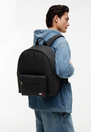 Joven con chaqueta vaquera y vaqueros, llevando una gran mochila negra de Tommy Jeans, ligeramente girado de lado sobre un fondo blanco.