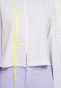 Lyse lilla ternet skjorte med gule dekorative snører, der har et cropped design og en diskret frontslids. Tekstureret stofdetalje er synlig.