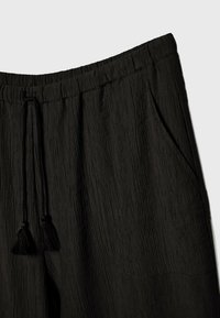 Zwarte, gestructureerde broek met een elastische tailleband, een trekkoord met kwastjes en zijzakken. De gerimpelde stof zorgt voor visuele interesse.