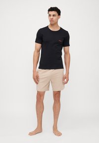 Svart bomull t-skjorte med rund hals og rødt logo, kombinert med beige shorts med et subtilt mønster. Modellen står barføtt.