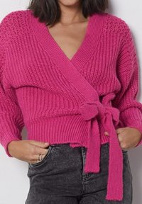 Rosa stickad omslagströja med djup V-ringning, ribbad textur, långa ärmar och knytning vid midjan. Bärs över grå jeans.