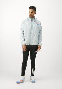 Hellgraue Zip-Jacke mit blauem und rotem Logo, kombiniert mit schwarzen, eng anliegenden Hosen mit weißen Streifen und hellblauen Fußballschuhen.