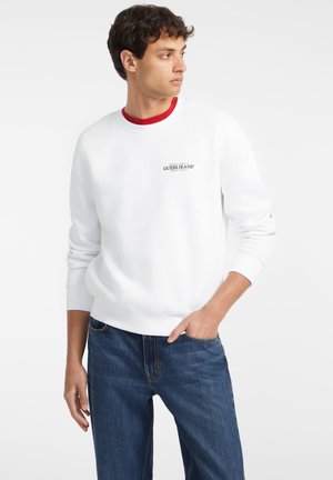 Sweatshirt blanc à col rond avec le logo "GUESS JEANS" en rouge et noir, porté sur un t-shirt rouge, associé à un jean en denim bleu.