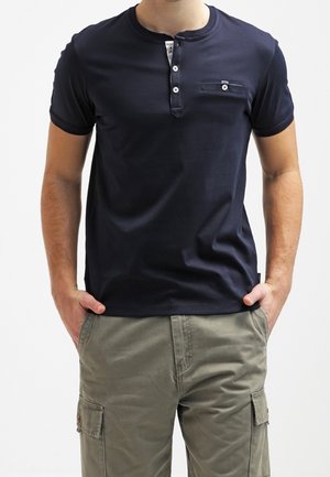 Mężczyzna ubrany w granatową koszulkę henley z krótkim rękawem i zapinanym na guziki kieszonką na piersi oraz oliwkowo-zielone spodnie typu cargo, z rękami w kieszeniach.