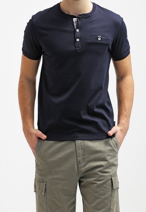 Homme portant un t-shirt henley bleu marine à manches courtes avec une poche poitrine à boutons et un pantalon cargo vert olive, les mains dans les poches.