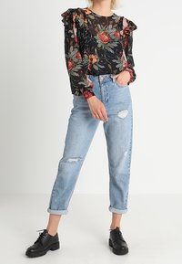Zwarte bloes met bloemenpatroon en geribbelde mouwen, gecombineerd met verouderde lichtblauwe jeans en zwarte veterschoenen. Casual, ontspannen pasvorm.