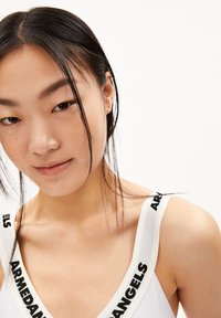 Witte tanktop met zwarte "ARMEDANGELS" opdruk op de schouderbanden. Het ontwerp heeft een brede halslijn en een zachte, gladde textuur. Close-up van het gezicht van het model.