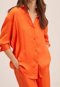 Oranžová, volná blůza vyrobená z hladké tkaniny, s límcem, zapínáním na knoflíky a ohrnutými manžetami. Kapsy na obou stranách.