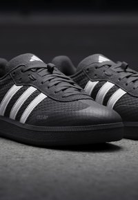 Czarne buty sportowe z teksturowaną powierzchnią, z trzema białymi paskami, okrągłym noskiem, sznurowanym zapięciem oraz logo po boku.