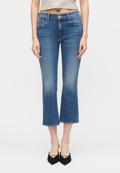 Γυναίκα που φοράει μπλε καμπάνα cropped τζιν, ριγέ cropped τοπ και μαύρες μυτερές γόβες, στέκεται μπροστά σε λευκό φόντο.