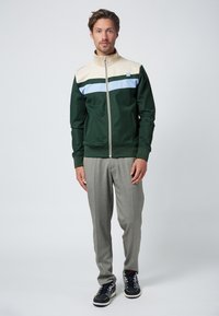 Mäns zip-up jacka i mörkgrön, med en krämfärgad överdel och blå rand. Matchad med ljusgrå byxor och navy atletiska skor.