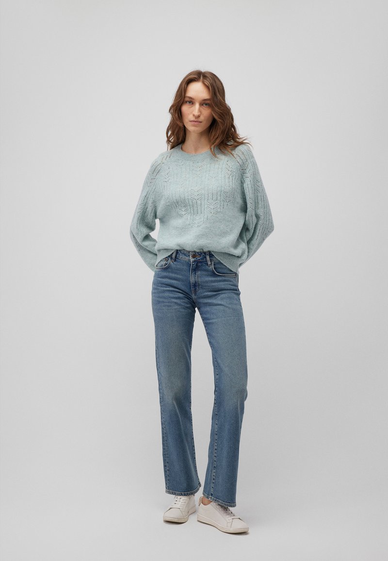 Hellblaues Strickoberteil mit strukturiertem Muster, kombiniert mit hochgeschnittenen blauen Jeans. Weiße Sneakers runden das Outfit ab.