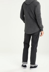 Dlouhý rukáv, tmavě šedá mikina s kapucí a žebrovanými detaily na rukávech, v kombinaci s tmavými džíny a černými teniskami s bílými akcenty.