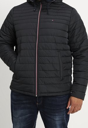 Veste noire matelassée avec coutures horizontales, fermeture éclair sur le devant, poches latérales et petit logo brodé sur la poitrine gauche, portée avec un jean foncé.