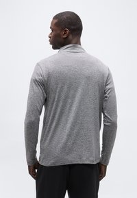 Άν δρας με την πλάτη, φορώντας ένα μακρυμάνικο, ανοιχτό γκρι πουκάμισο με λεπτές ρίγες και μαύρο παντελόνι, μπροστά σε απλό λευκό φόντο.