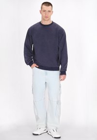 Felpa navy a maniche lunghe, abbinata a pantaloni cargo azzurri con tasche laterali e sneakers bianche con rifiniture nere.