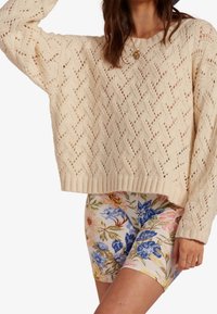 Cremfarbener Strickpullover mit einem strukturierten Rautenmuster, kombiniert mit floralen Radshorts in Pastellfarben.