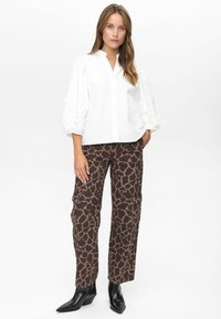 Vrouw in een witte blouse met pofmouwen, bruine cargobroek met giraffeprint en zwarte puntige enkellaarzen, staand met één hand in haar zak.