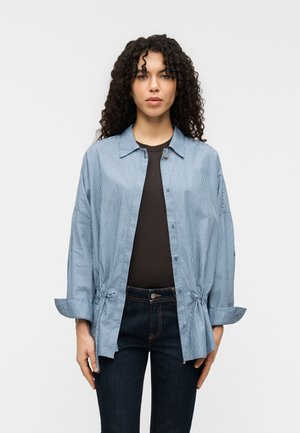 Blau-weiß gestreiftes Hemd mit Knöpfen, Kragen, weitem Schnitt und hochgekrempelten Ärmeln, kombiniert mit einem schwarzen Oberteil und dunklen Jeans.