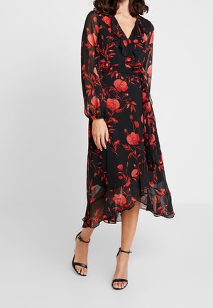 Kvinde iført en sort gennemsigtig midi-kjole med rødt blomsterprint, udskåret V-hals, lange ærmer og sorte hælede sandaler med ankelrem.