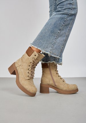 Bottines en suède beige avec un talon empilé, un laçage à l'avant et une fermeture éclair sur le côté. Présente un accent marron au niveau du haut et une semelle texturée.