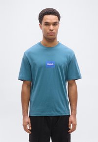 Άνδρας που στέκεται με το πρόσωπο προς τα εμπρός, φοράει μια τιρκουάζ μπλούζα με μια μπλε τετράγωνη ετικέτα στο στήθος που γράφει "HUGO" και μαύρο παντελόνι.