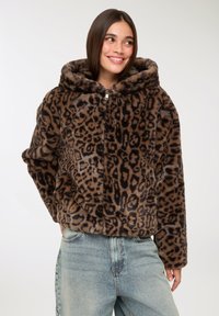 Donna che indossa un giubbotto con cappuccio in pelliccia sintetica stampata leopardata marrone e nera, con jeans a vita alta azzurri chiari, sorridente e con uno sguardo di lato.
