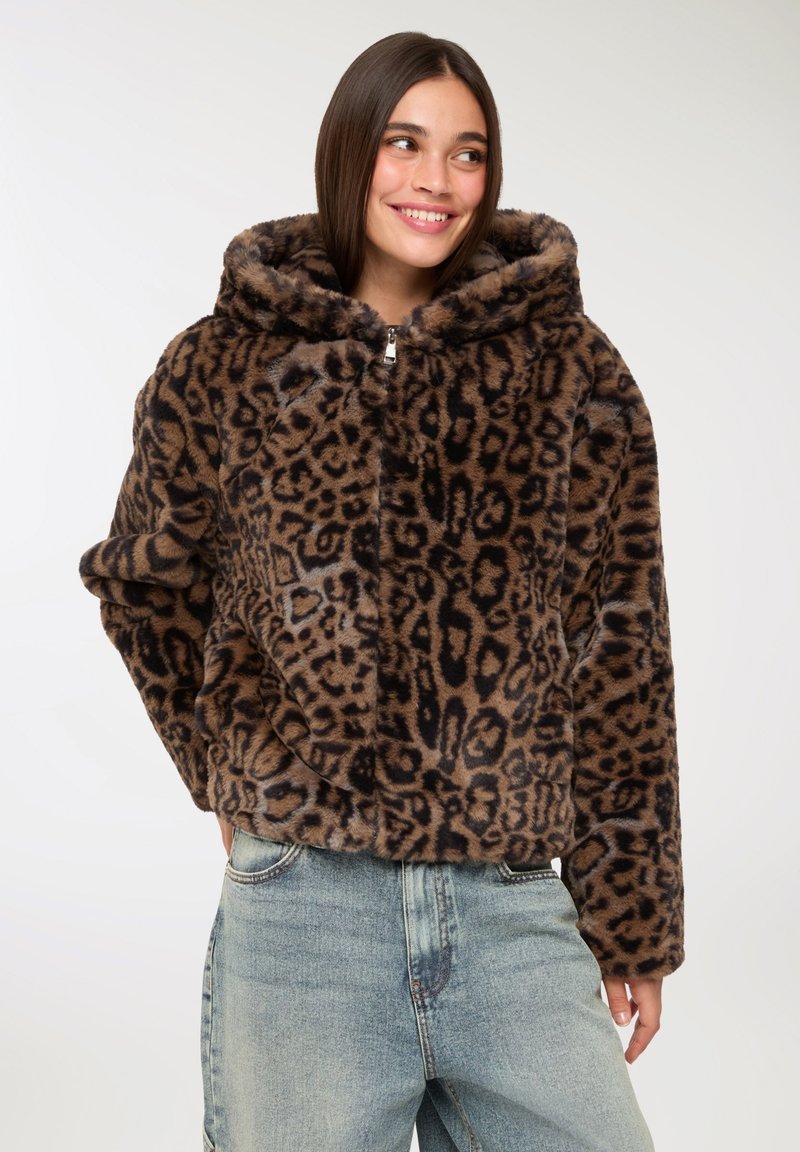 Donna che indossa un giubbotto con cappuccio in pelliccia sintetica stampata leopardata marrone e nera, con jeans a vita alta azzurri chiari, sorridente e con uno sguardo di lato.