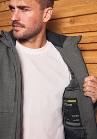 Grå isolerad jacka med en texturerad yta, med huva, dragkedja fram och en innerficka märkt "äventyr". Bärs över en vit t-shirt.