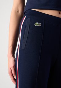 Marineblaue Jogginghosen mit roten und weißen Seitenstreifen, aus weichem Stoff. Verfügt über ein kleines gesticktes Krokodil-Logo und schräge Taschen.