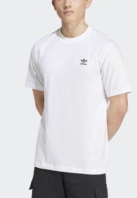 Bílá bavlněná tričko s krátkým rukávem, kulatým výstřihem a malým černým logem Adidas na levé části hrudi. Hladká textura, volný střih.