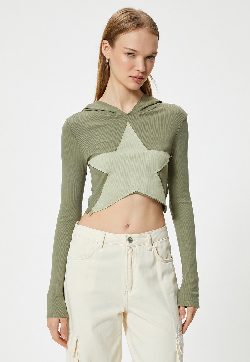Sudadera corta de color verde oliva con un gran diseño de estrella en un tono verde claro. Presenta mangas largas y un dobladillo sin rematar, combinada con pantalones de color crema.