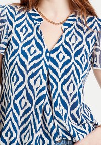 Short-sleeve blouse in white and blue with a diamond pattern, featuring a V-neckline and smooth, lightweight fabric.