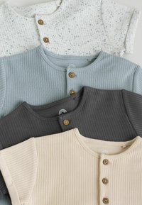 Cuatro camisetas de bebé de manga corta y tejido acanalado, dobladas, en colores crema, gris oscuro, azul claro y blanco, con botones de madera, sobre un fondo blanco.