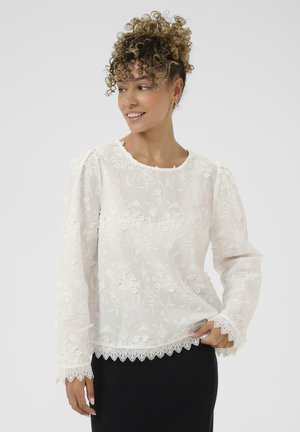 Blouse blanche brodée avec des motifs floraux, texture transparente, encolure ronde et bordure en dentelle festonnée sur les manches et l'ourlet. Manches longues.