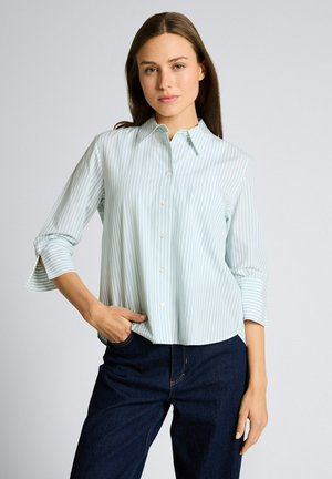 Femme portant une chemise à rayures bleu clair et blanche à boutons avec manches trois quarts et un jean bleu foncé, posant avec une main dans la poche.