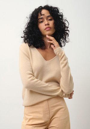Beige trui van zacht materiaal met een v-hals en lange mouwen, gecombineerd met lichtbeige broek. De trui heeft een losse pasvorm en een zachte textuur.
