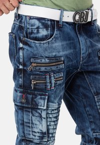 Dunkelblaue Denim-Cargohosen mit verblasster Textur. Verfügt über mehrere mit Reißverschlüssen verschlossene Taschen, rote Nähte und einen weißen Gürtel.