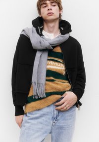 Fekete, cipzáros dzseki, oversized fazonnal, egy csíkos barna, zöld és sárga bolyhos kötött pulóver fölé rétegezve. Szürke sál lazán lógva. Világoskék farmernadrág.