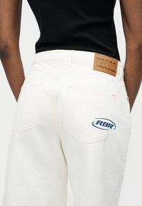 Osoba nosząca białe dżinsy z skórzaną naszywką Red Bull Racing x Pepe Jeans oraz niebieskim logo RBR na tylnej kieszonce, zestawione z czarnym topem.