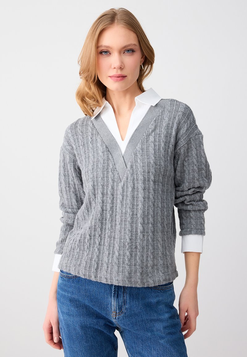 Femme blonde portant un pull à col en V gris à torsades par-dessus une chemise blanche à col, associée à un jean bleu, debout devant un fond clair.